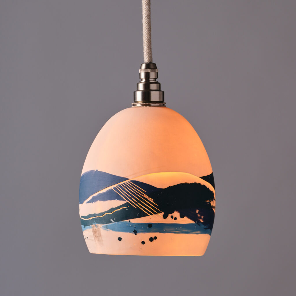 Adur Art Collective artwork by member artist Amy Frankie Smith, a lit pendant ceramic lampshade titled Sea Lamp in peach with abstract patterns in blue hangs down against a grey background.
